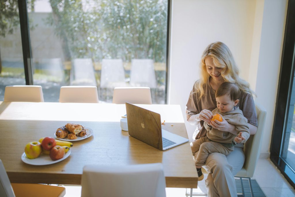 Blonde mother holding baby with laptop at dining table, enjoying breakfast indoors.